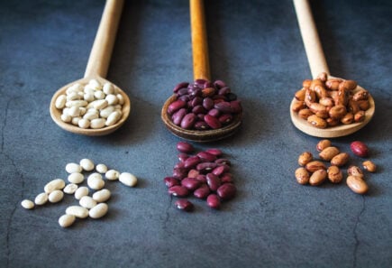 Three wooden spoons holding red, brown, and white beans on a dark gray background