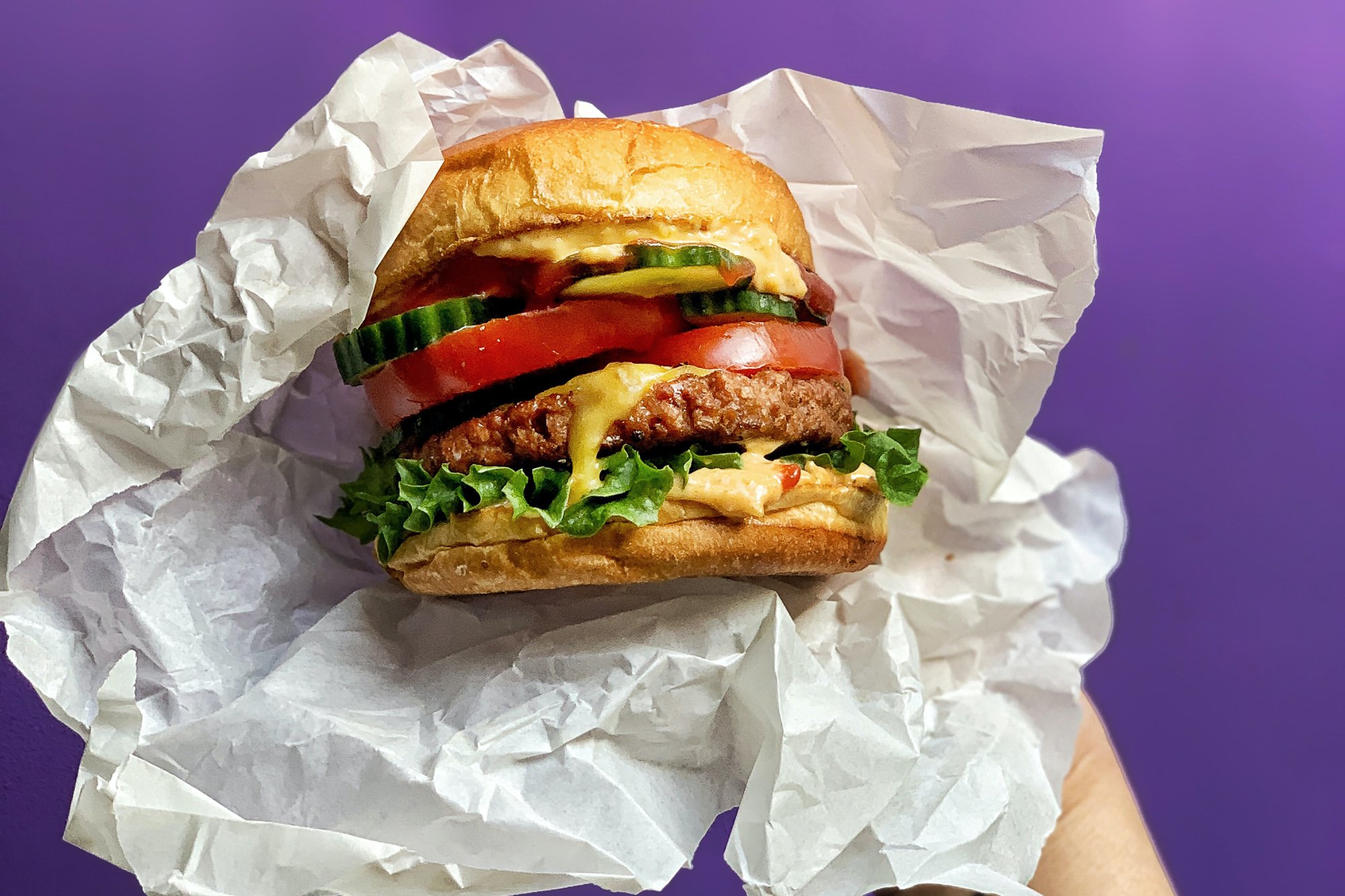 LikeMeat burger in white paper being held in front of a purple background