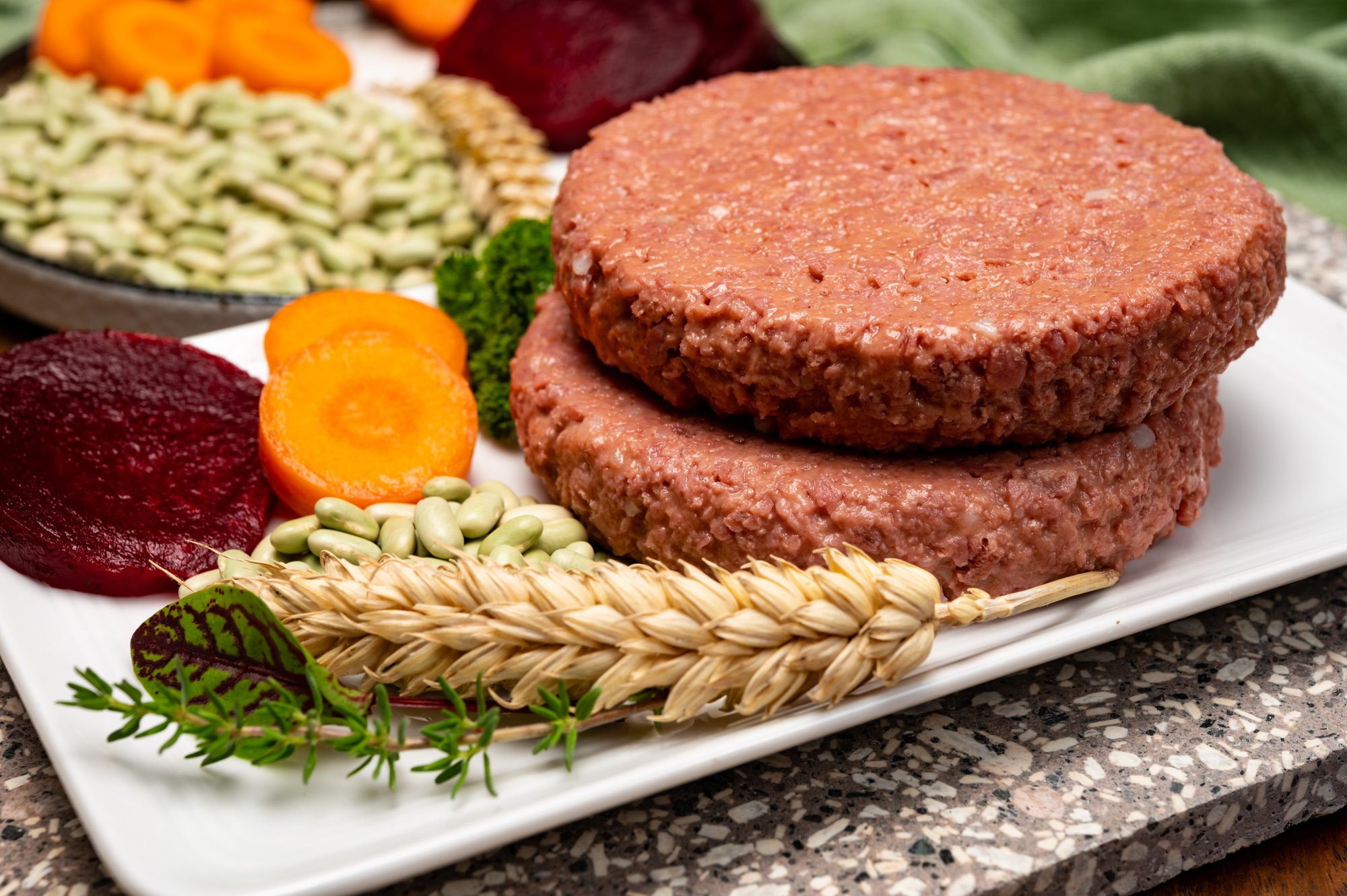 Plant based soy protein burger patties on a white plate next to wheat, legumes, carrots and beets