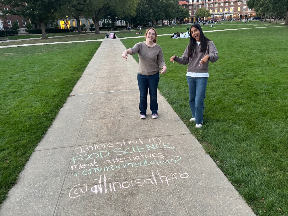 Two members of the UIUC Alt Protein Project standing beside chalk messages about alternative proteins on the campus quad.
