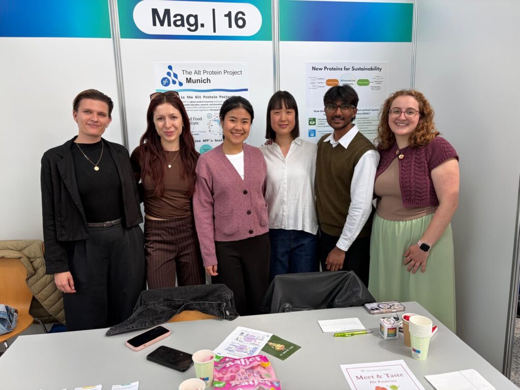 Six members of the Munich Alt Protein Project executive team and their GFI mentor standing at a table during the TUM Sustainability Fair.