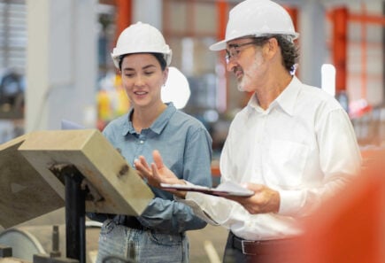 Two engineers wearing hard hats review documents and discuss a project inside an industrial facility.